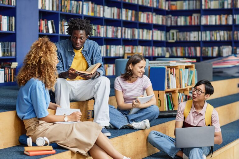 Quatro estudantes de diferentes etnias sentados em degraus de uma biblioteca moderna, conversando e estudando com livros e um notebook.